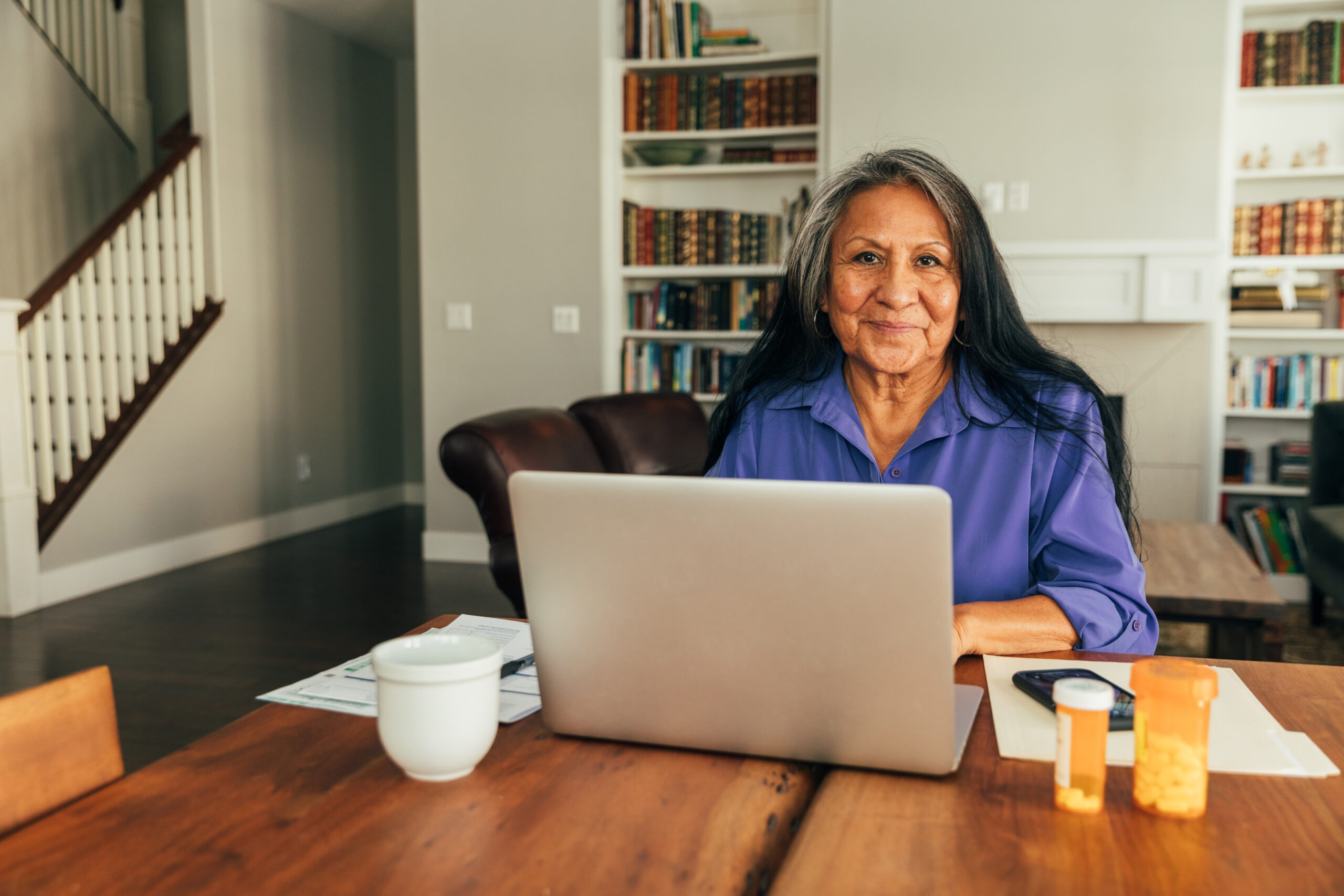 A senior Indigenous woman sits at her kitchen table while paying medical bills, talking with her doctor, and updating medicine prescriptions.