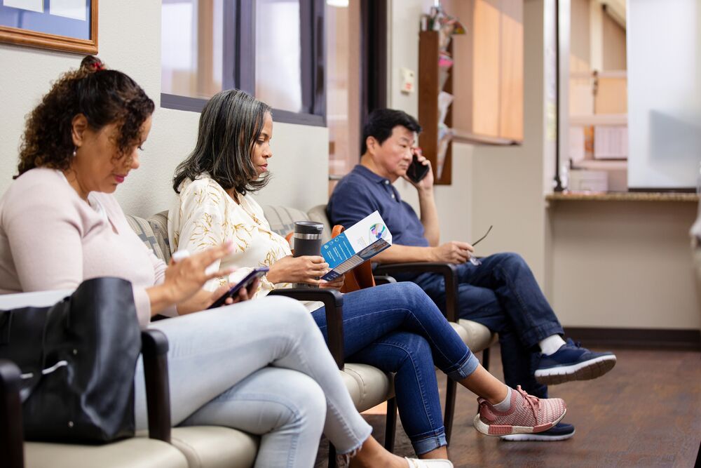 3 patients sit in a waiting room. 2 use a cell phone to talk and text and one reads a pamphlet.