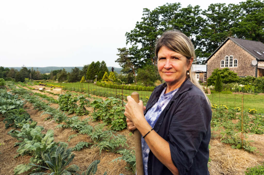 A woman stands outside with a crop of growing vegetables. She holds a tool and looks at the camera.