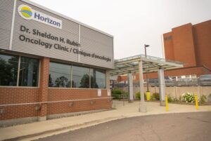 The exterior of the Dr. Sheldon H. Rubin Oncology Clinic in Moncton, New Brunswick. The building features a brick and metal facade with a sign reading 'Horizon Health Network' and the clinic's name in both English and French. A glass-covered entrance walkway is visible, along with nearby parking and landscaping.