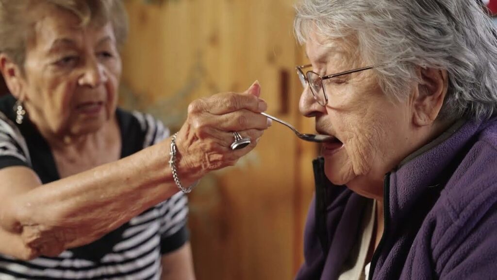 Deux femmes âgées membres des Premières Nations : celle de gauche nourrit celle de droite à la cuillère.